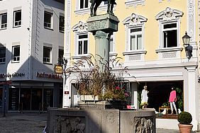 Auf einem weiten Platz steht ein steinerner Brunnen mit Figuren an den Seiten. In der Mitte erhebt sich eine Säule mit einem Reiter mit Pferd aus Bronze. Der Reiter bläst in ein Horn.
