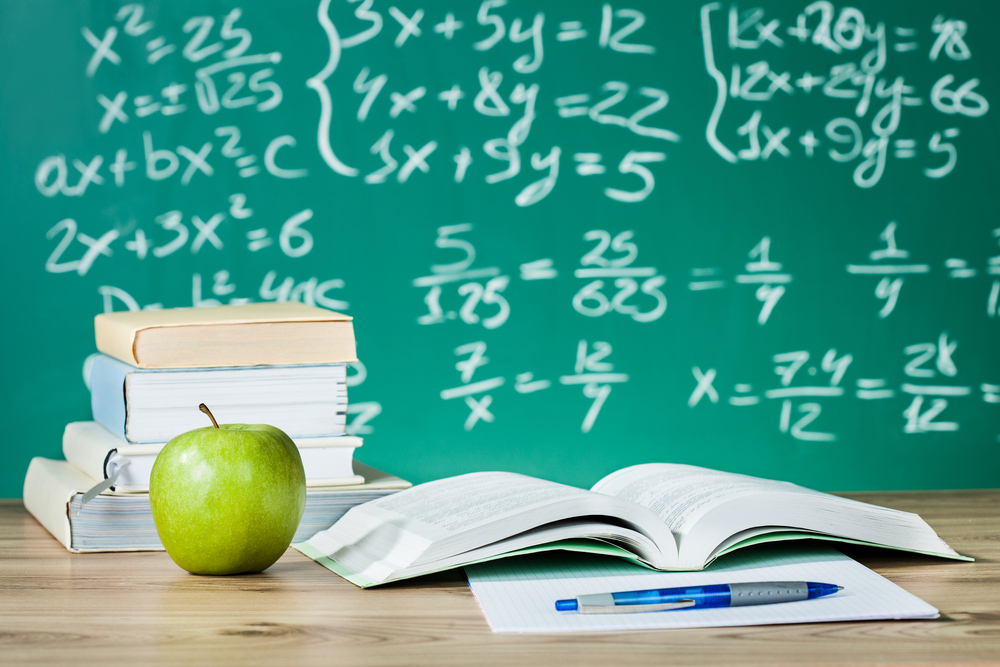 Auf einem Tisch, vor einer Tafel mit mathematischen Formeln, liegen Bücher und ein Apfel.