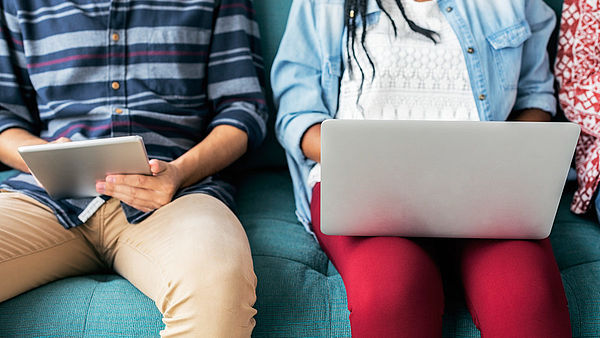 A student with a laptop on her lap and a student with a tablet in her hand are sitting on the sofa.