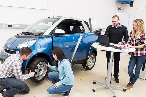 A blue Smart car with two students kneeling in front of it on the left. Two more students are standing at a table in front of a laptop.