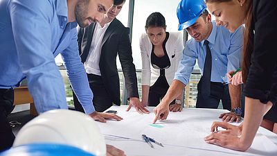 Eine Gruppe von Ingenieurinnen und Ingenieuren sowie anderen Bauplanern stehen um einen Tisch und betrachten einen Bauplan.