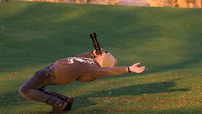 Ein Fotograf auf einer Wiese lehnt sich weit in Rückenlage und hält die Kamera in Richtung Himmel.