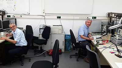 Ein Raum in dem links und rechts jeweils ein Professor vor einem Schreibtisch sitzt. 