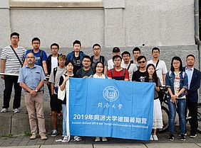 Eine Gruppe chinesischer Gäst mit einem deutschen Professor und einer blauen Flagge vor einem Hochschulgebäude.