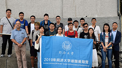 Eine Gruppe chinesischer Gäste mit einem deutschen Professor und einer blauen Flagge vor einem Hochschulgebäude.