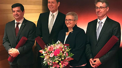 Drei Männer und eine Frau vor einer roten Wand. Die Frau hält einen Blumenstrauß.