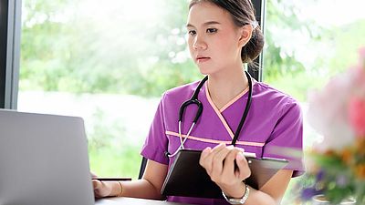 Eine Pflegerin steht mit einem Klemmbrett vor einem Laptop.