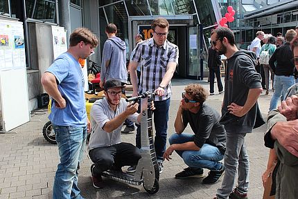 Studierende und Besucher betrachten ein selbst gebautes Elektro-Gefährt