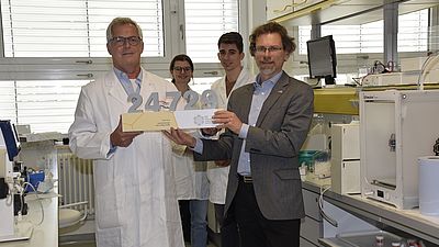 Zwei Männer bei einer Preisübergabe in einem Labor - zwei Studierende im Hintergrund