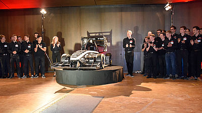 Auf einer Bühne steht in der Mitte ein Rennauto. Links und rechts daneben stehen mehrere Menschen.
