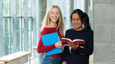 Zwei Studentinnen stehen auf einem Gang und halten Hefte und Bücher.