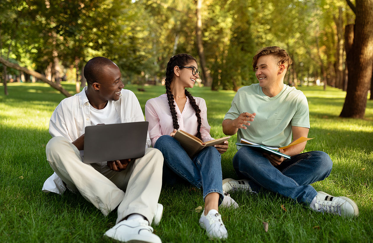 Gruppe internationale Studierende Gruppe internationale Studierende sitzt im Außenbereich des Campus mit Laptop und Arbeitsbüchern Happy international students resting in college campus, sitting on grass, talking and using laptop computer. Friends preparing for classes or exams outdoors